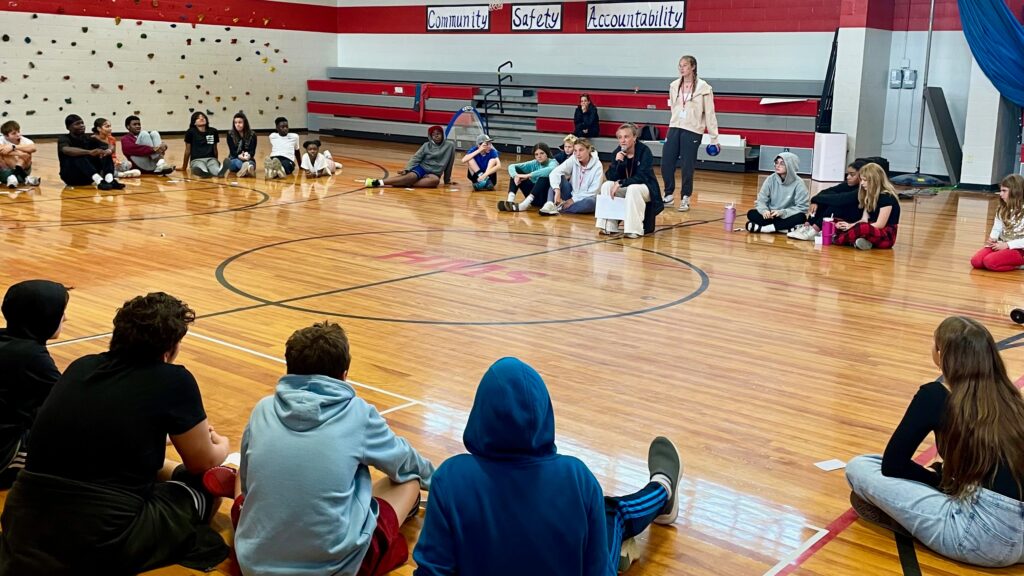 Students seated in a circle, listening to a speaker holding a microphone.